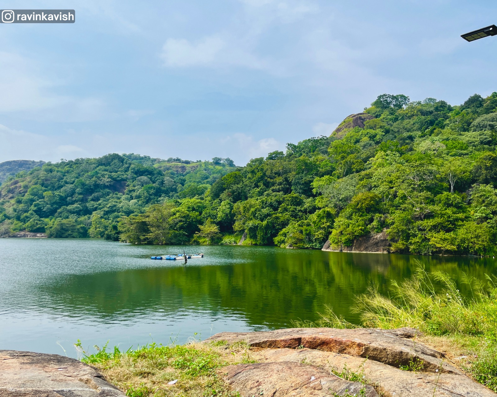 Buduruwagala Reservoir in Ella with calm waters, tree-covered hills in the background, and a rock formation in the foreground showcasing Sri Lankas scenic landscapes