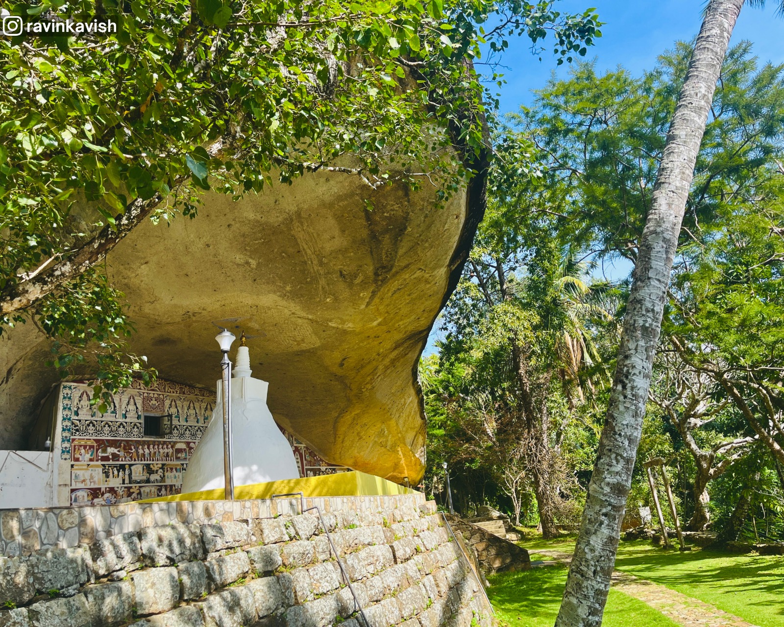 Cave temple rock shelter at Rakkiththa Kanda Rajamaha Viharaya housing a small stupa, ancient murals and statues, with a glimpse of the surrounding hills and greenery