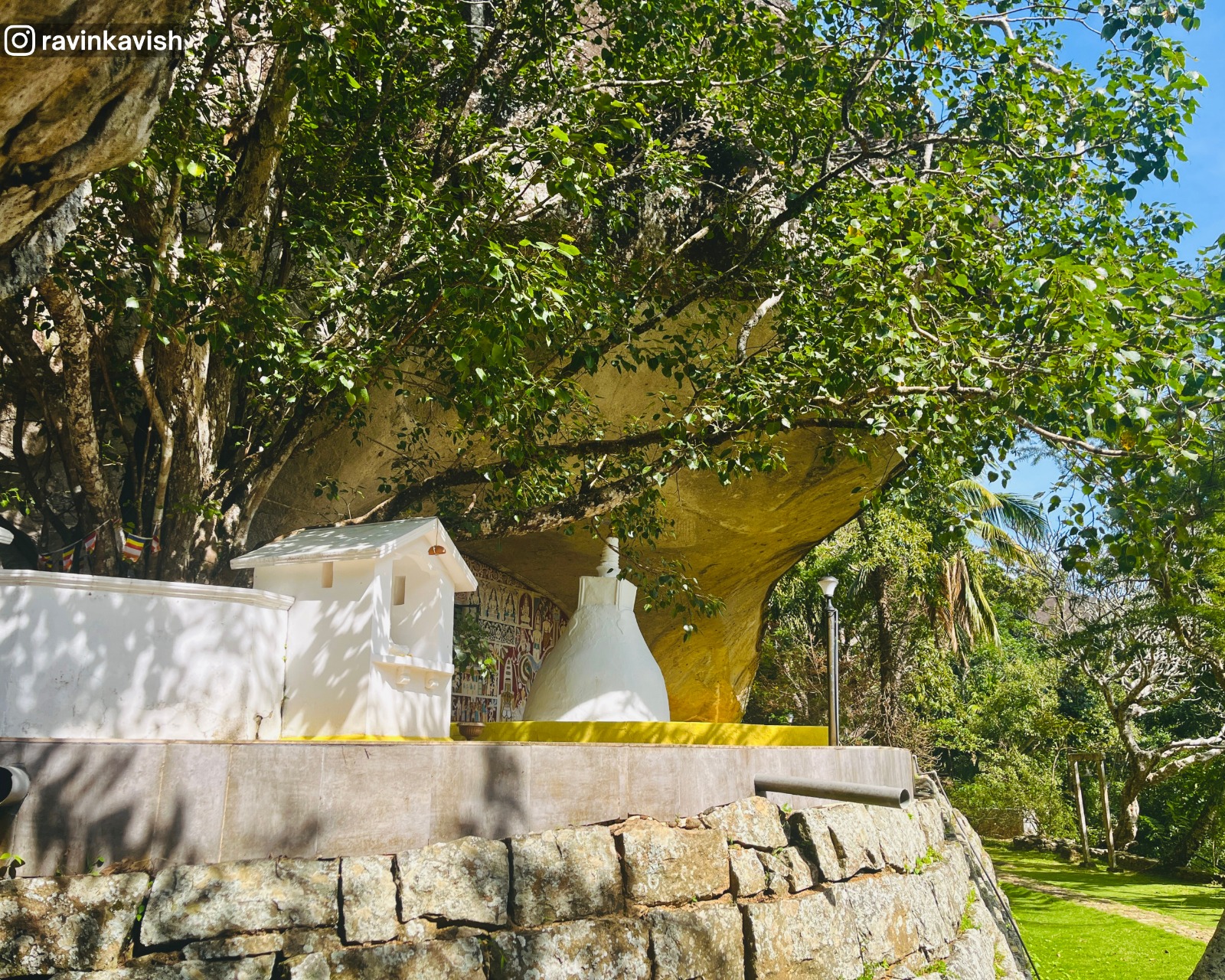 Cave temple rock shelter at Rakkiththa Kanda Rajamaha Viharaya with a small stupa, ancient murals and statues, and the sacred Bo tree along with surrounding hills and greenery
