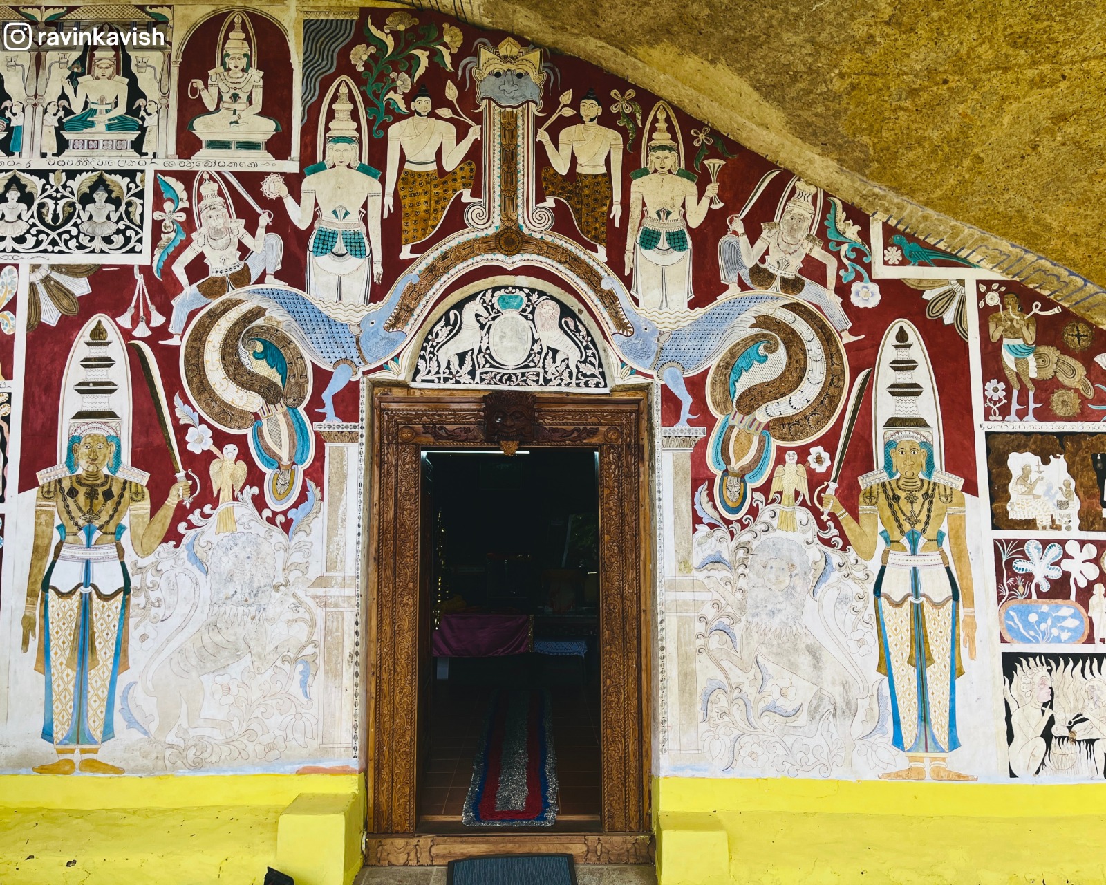 Close-up of the cave entrance at Rakkiththa Kanda Rajamaha Viharaya showing the door frame and surrounding murals, including colonial-era symbols and the Dragon Arch