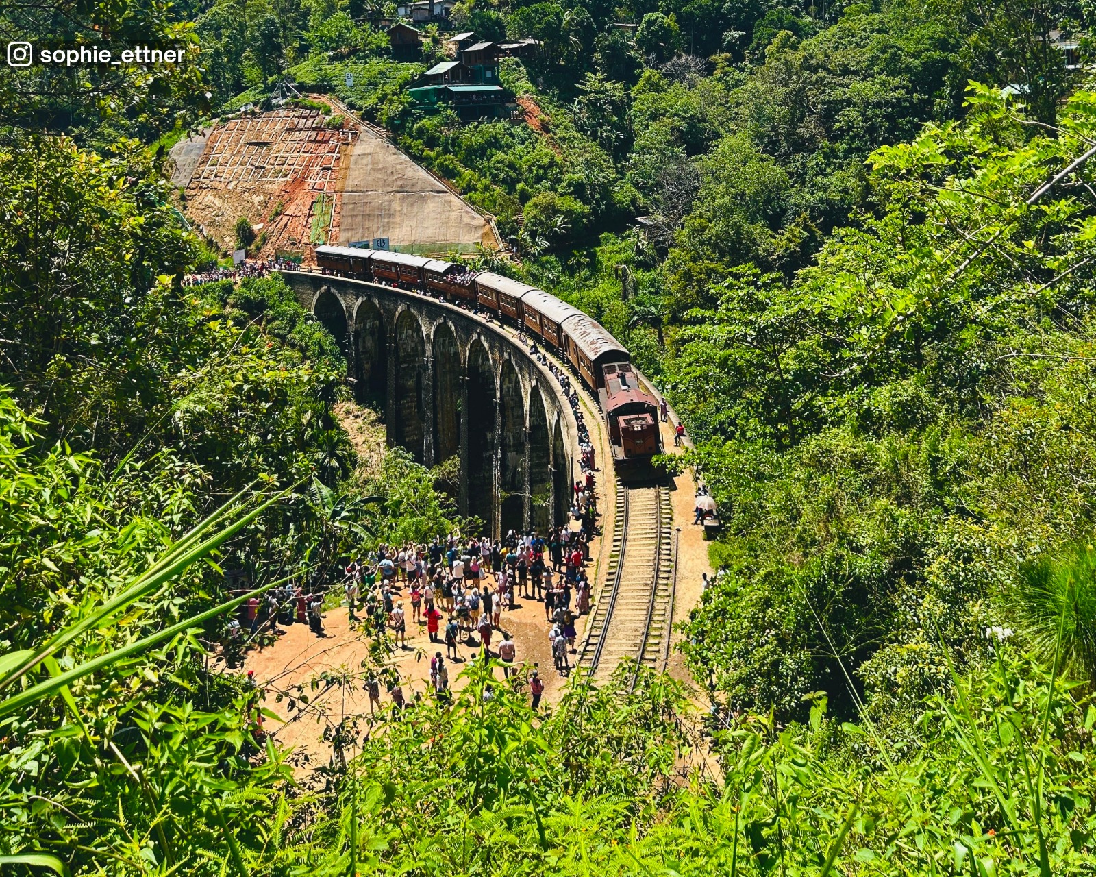 High-angle view of the Nine Arch Bridge near the tunnel entrance, showing a train traveling along the stone railway bridge amid lush greenery