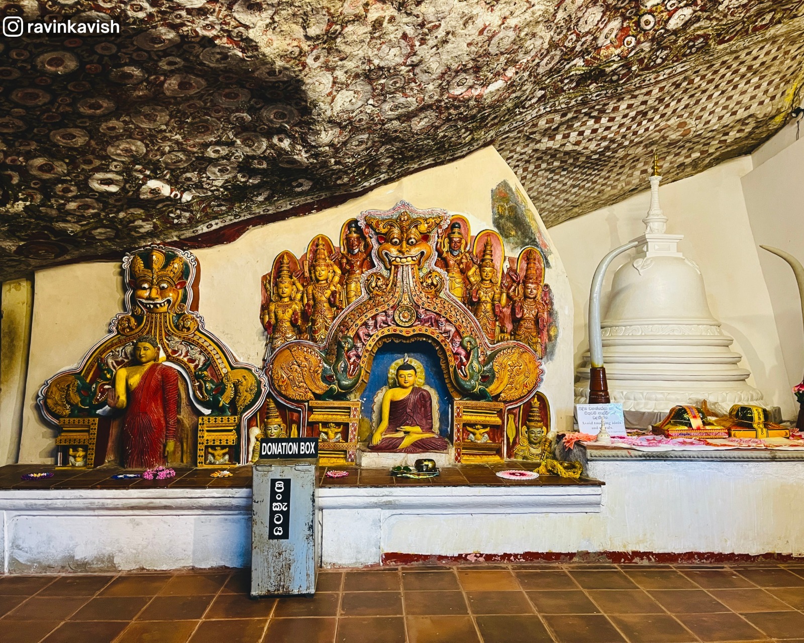 Interior of Ravana Royal Cave Temple near Ella, Sri Lanka, with a central seated Buddha under a dragon arch, surrounding god figures
