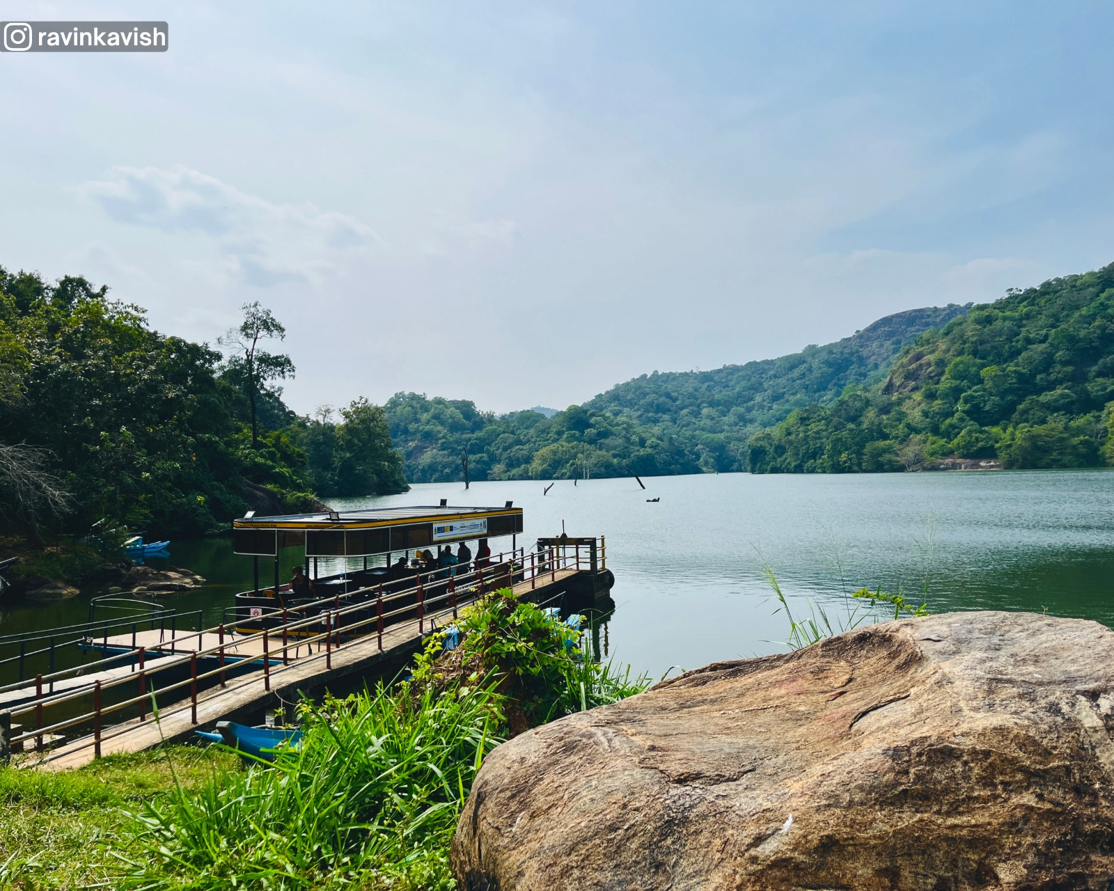 Jetty at Buduruwagala Reservoir in Ella with a small ferry, a large boulder on the bank, and surrounding tree-covered hills showcasing Sri Lankas scenic landscapes