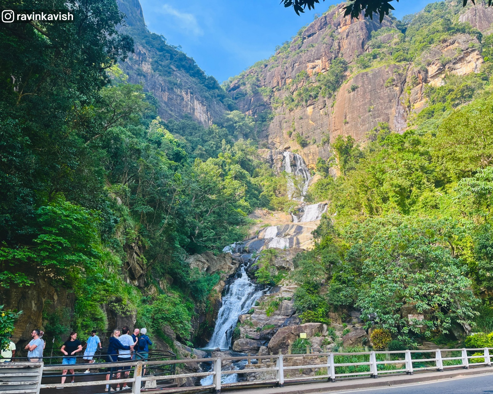 Ravana Waterfall viewed from across the road near Ella, Sri Lanka, showing the cascading waterfall, surrounding cliffs, and a small section of the road in the foreground