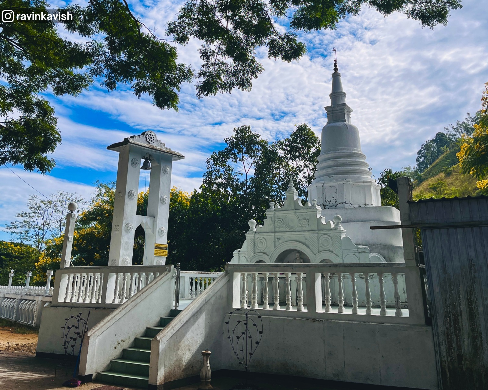 Stupa at Dowa Ancient Rock Temple with surrounding structures including the bell, flower offering area, staircase, and greenery with a view of the sky