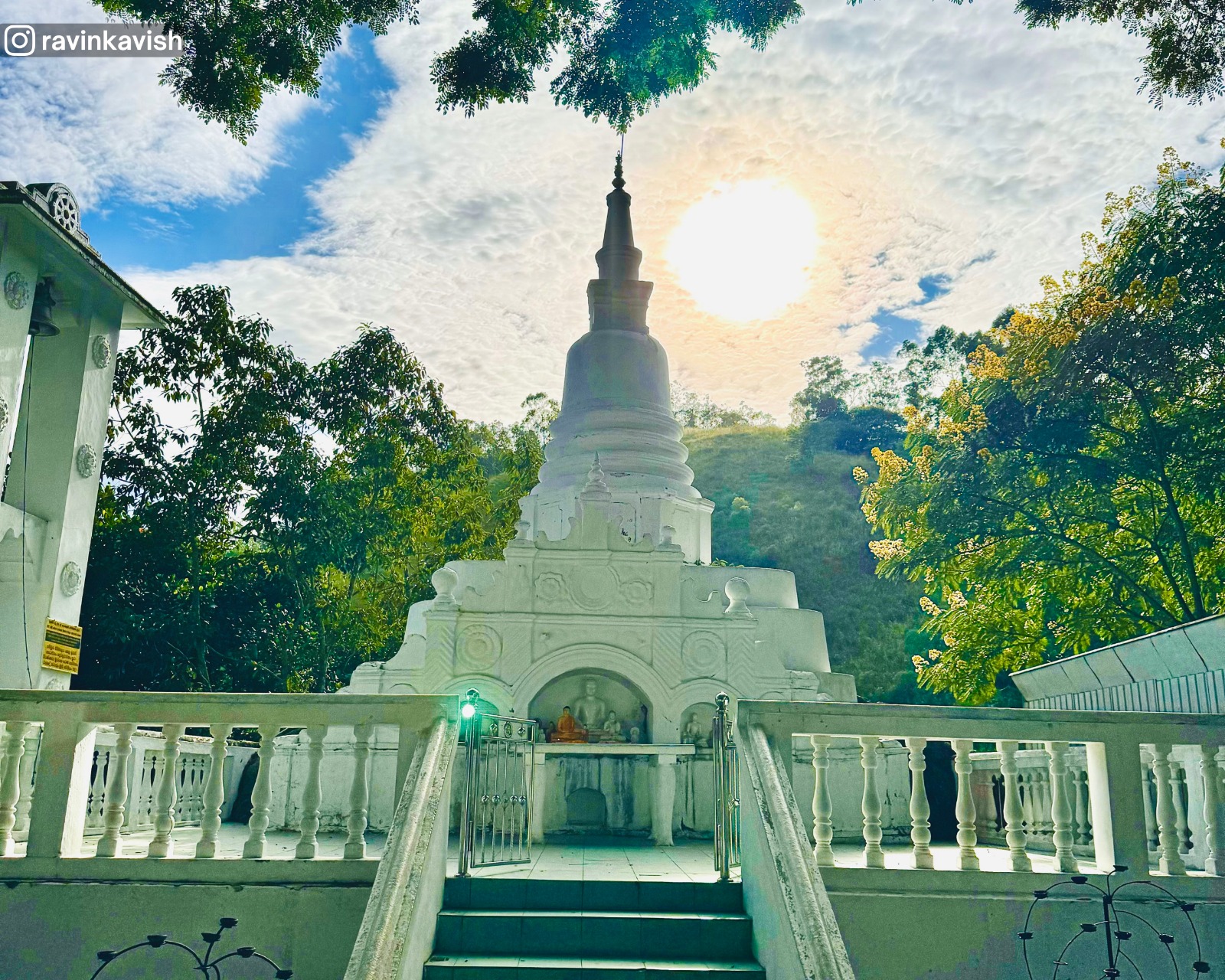Stupa at Dowa Ancient Rock Temple with the sun shining behind it, surrounded by the bell, flower offering area, staircase, and greenery