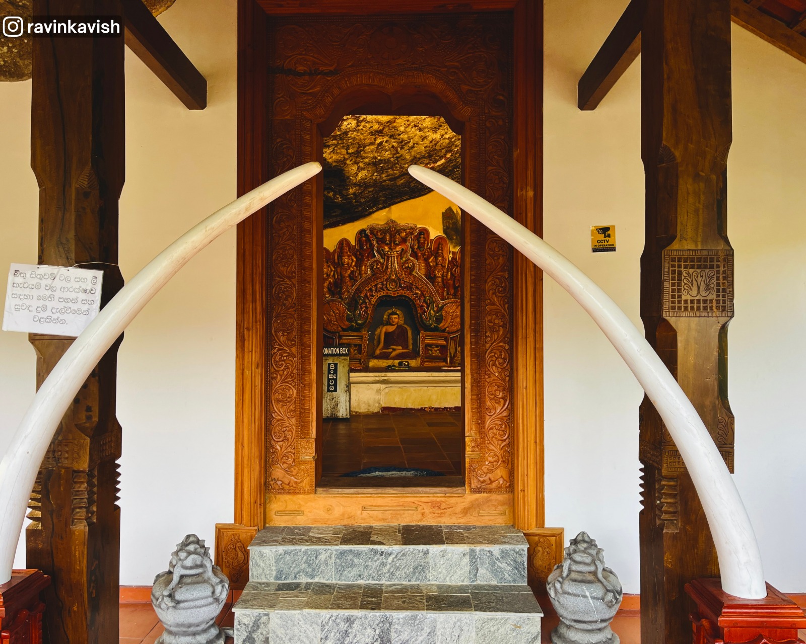 View through the doorway of Ravana Royal Cave Temple image house in Ella, Sri Lanka, featuring a seated Buddha, dragon arch, two roof pillars, and elephant tusks