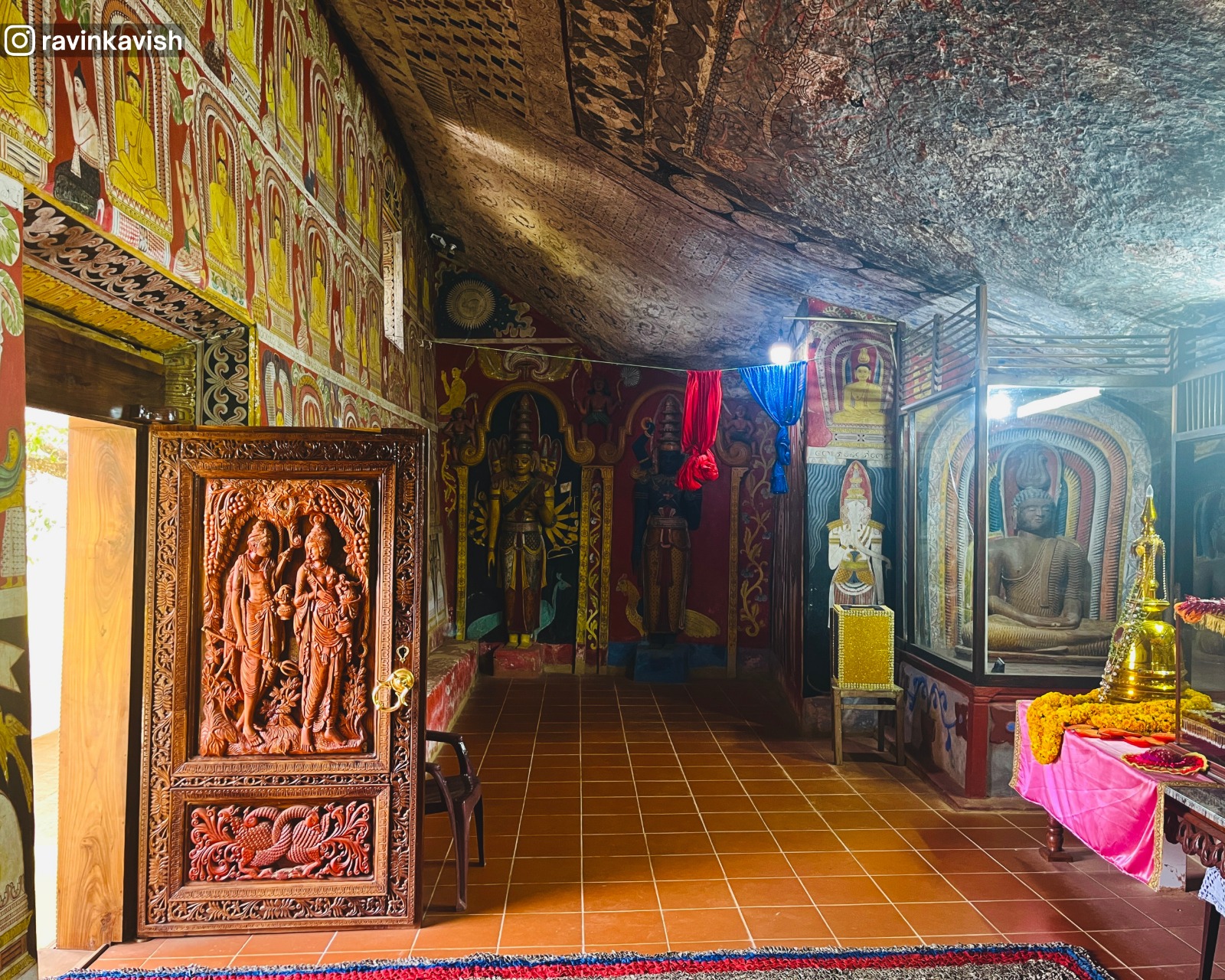 Wide view inside the cave at Rakkiththa Kanda Rajamaha Viharaya capturing multiple statues, wall murals, and the cave entrance door in a single frame
