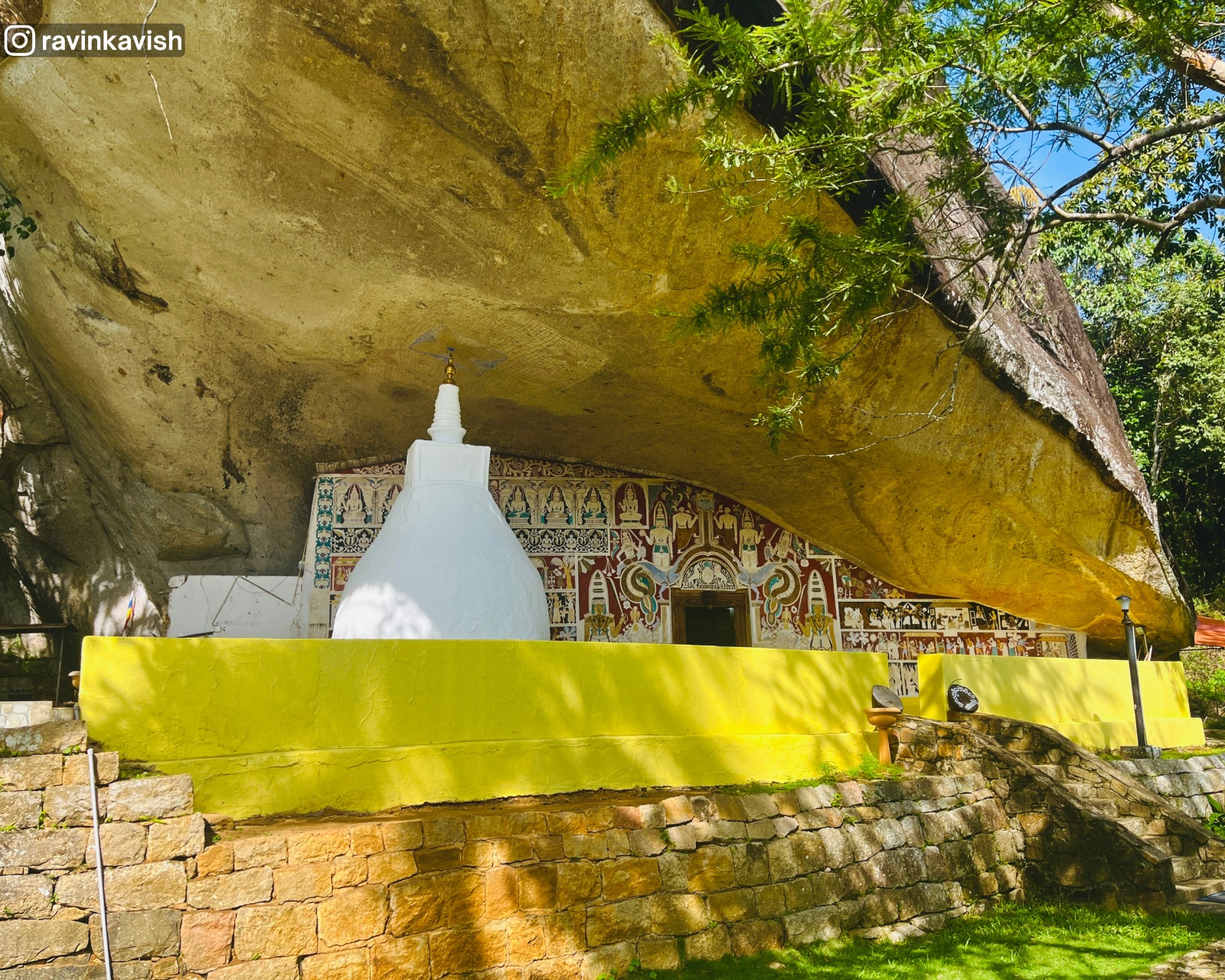 Wider view of the cave temple at Rakkiththa Kanda Rajamaha Viharaya showing the natural rock roof, small stupa, mural-covered inner wall, yellow outer wall, and stone staircase, with surrounding greenery