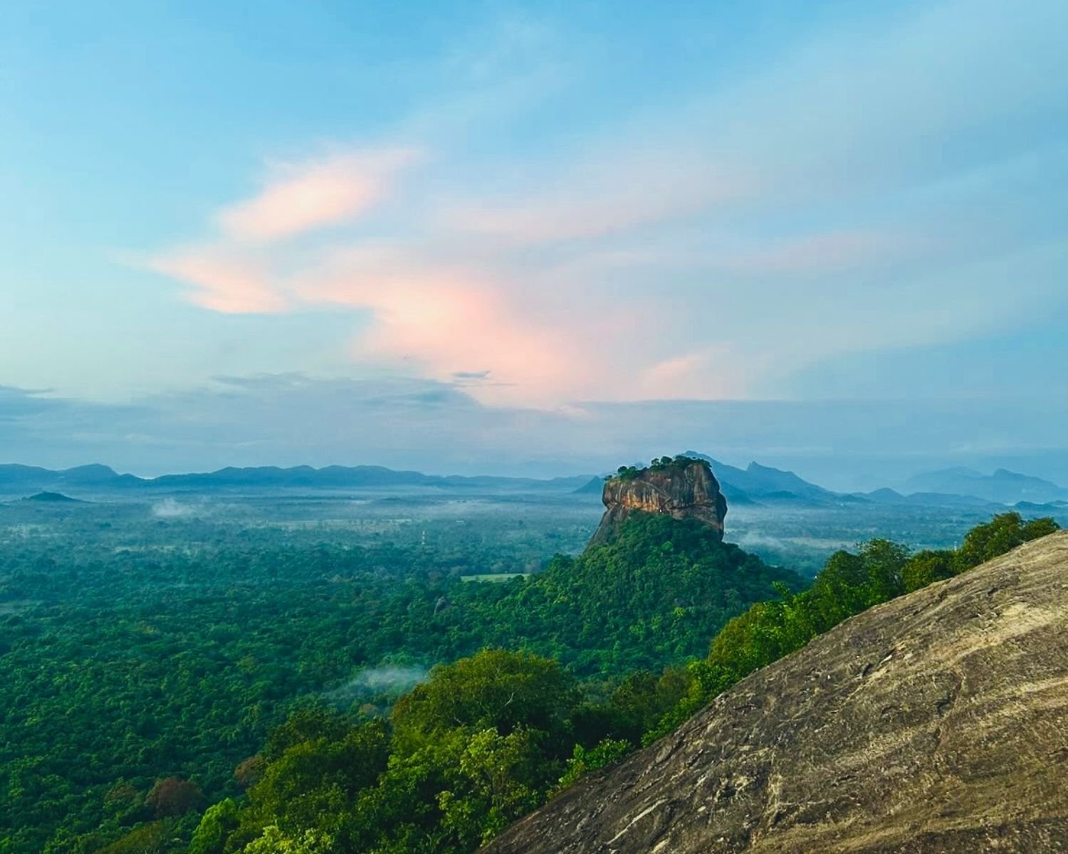 Early morning wide view of Lion Rock, the Sigiriya rock fortress, above misty jungle with pastel clouds and distant mountains, seen from rocky slope of Pidurangala near Sigiriya
