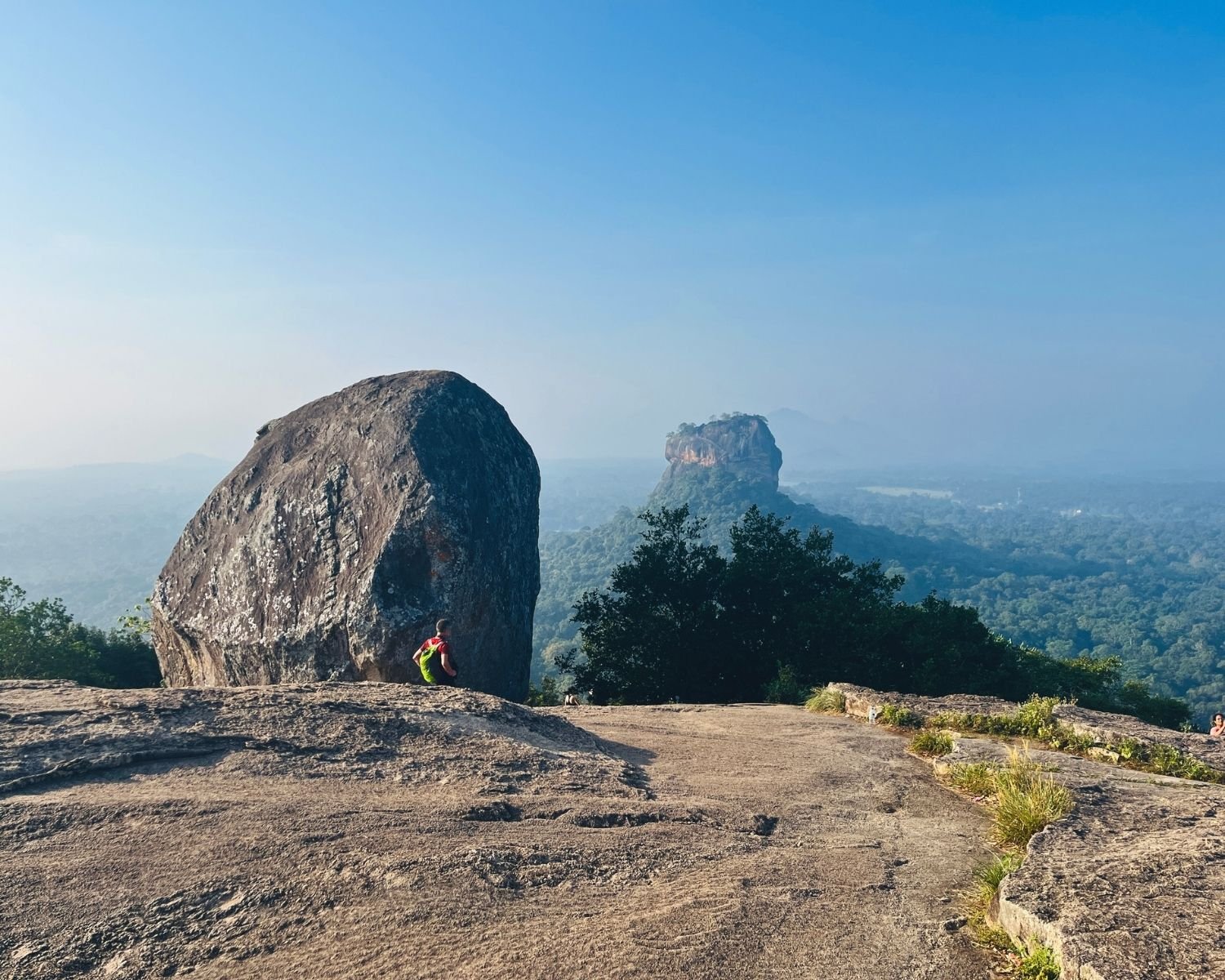 Large rounded boulder on rocky summit plateau with distant view of rock fortress above jungle canopy under clear blue sky, Pidurangala Rock near Sigiriya