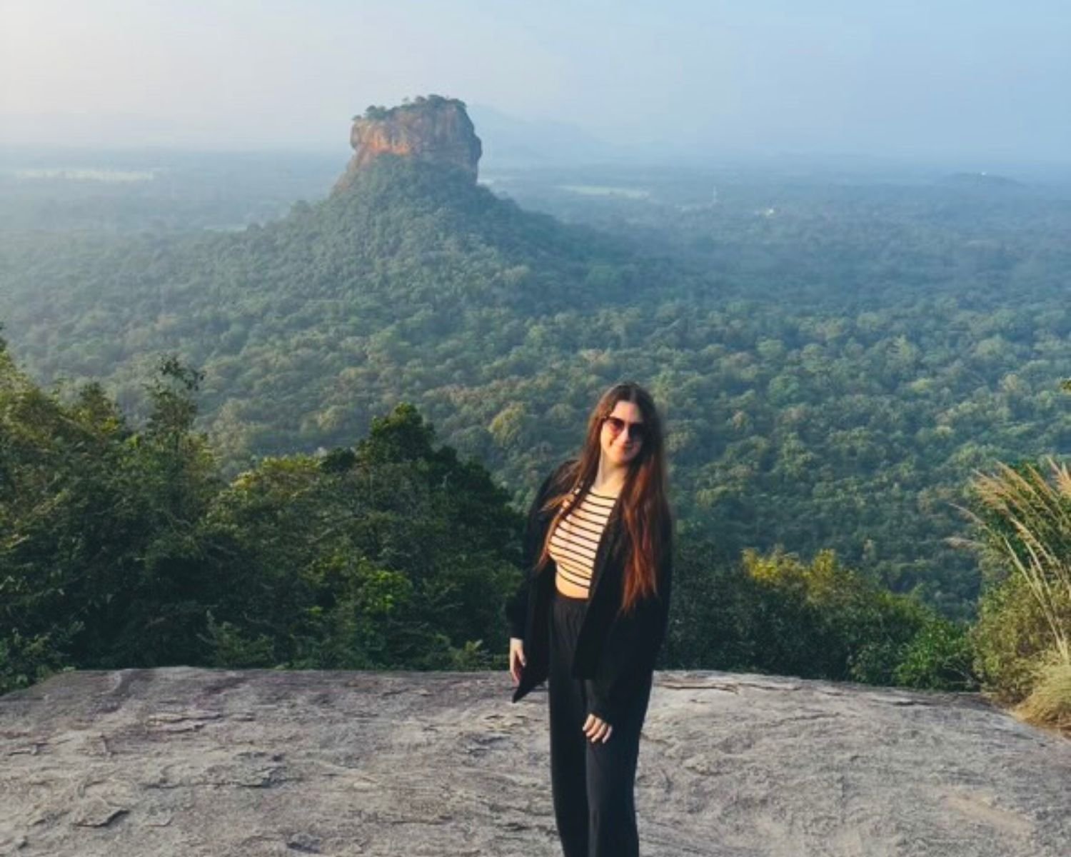 Woman standing on Pidurangala Rock summit in early morning with Sigiriya Rock Lion Rock rising from jungle in background, Sri Lanka