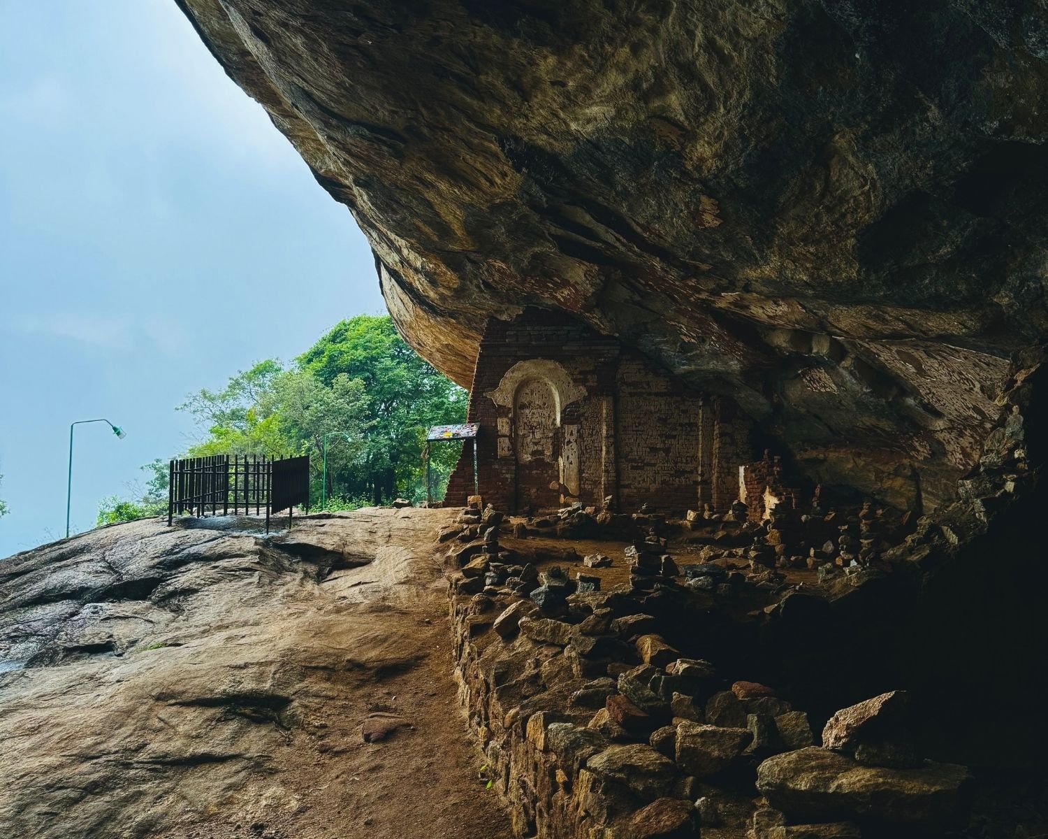 Ruined brick wall with faded arched alcove and stacked stone cairns under massive rock overhang halfway up Pidurangala Rock, located just before the cave temple with reclined Buddha statue, near Sigiriya