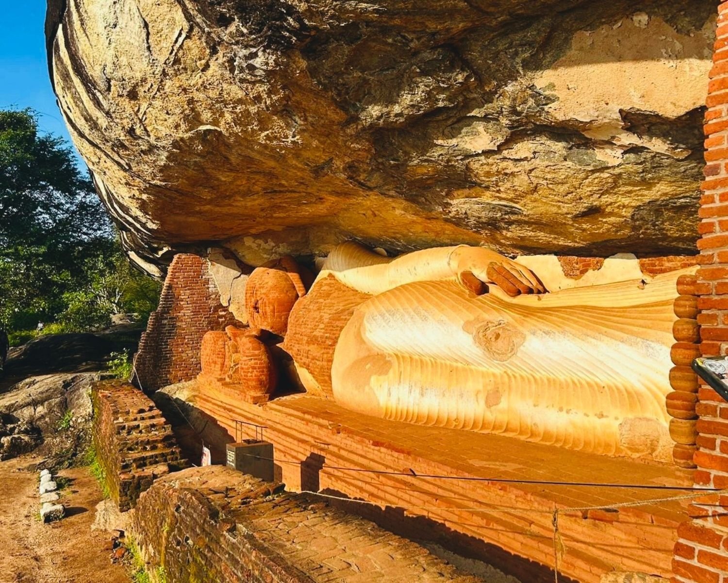Side view of reclining Buddha statue under massive rock overhang with brick walls and path, Pidurangala cave shrine near Sigiriya