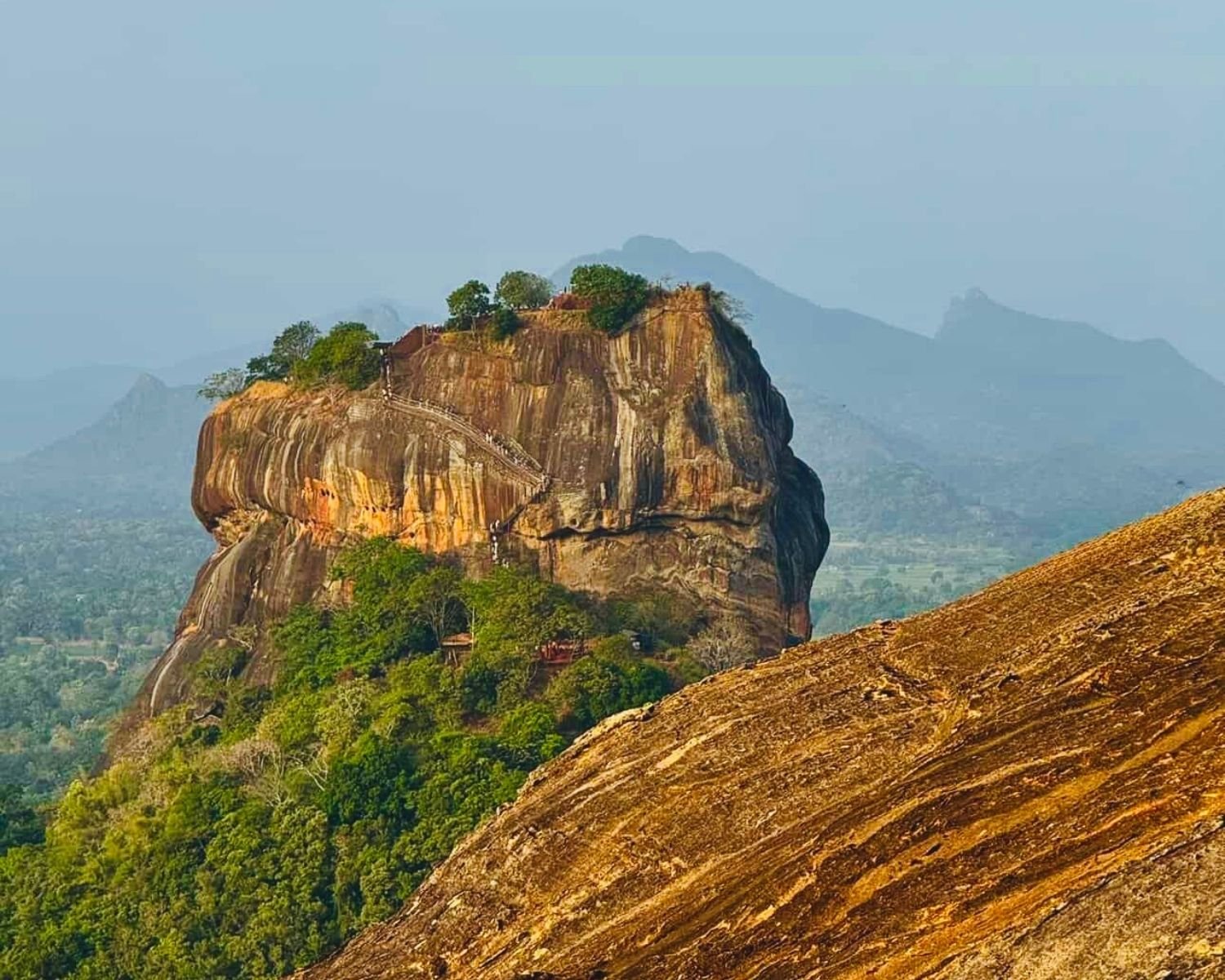 Close telephoto view of rock fortress with visible metal staircases on cliff face rising above jungle, seen from rocky slope of Pidurangala near Sigiriya