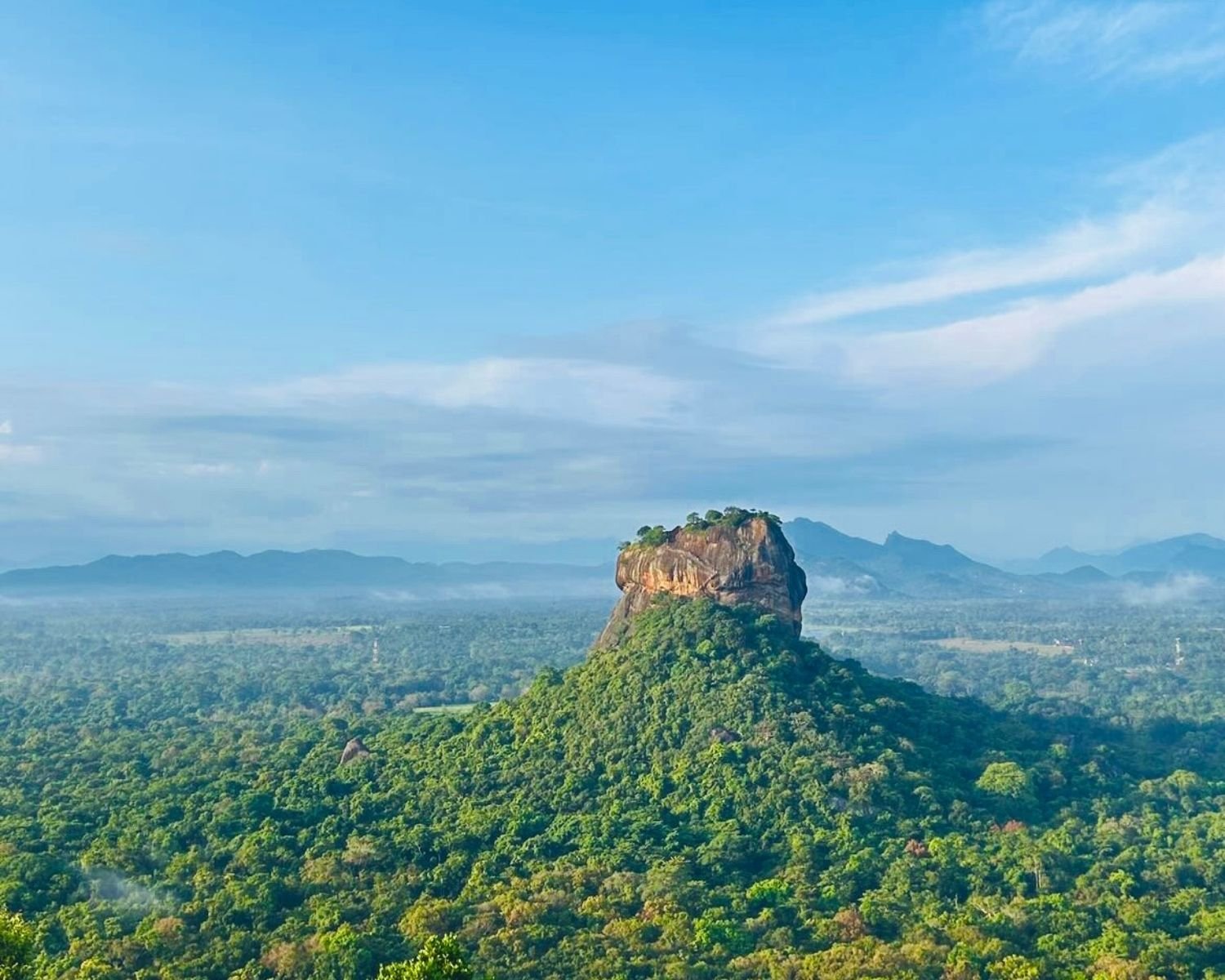 Sigiriya Lion Rock rising above dense jungle with blue mountains and morning mist, view from Pidurangala Rock near Sigiriya