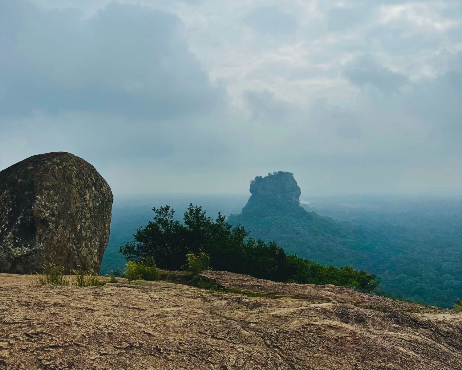 Distant view of Sigiriya rock fortress beyond jungle under cloudy sky, seen from rocky summit with large boulder at Pidurangala Rock near Sigiriya