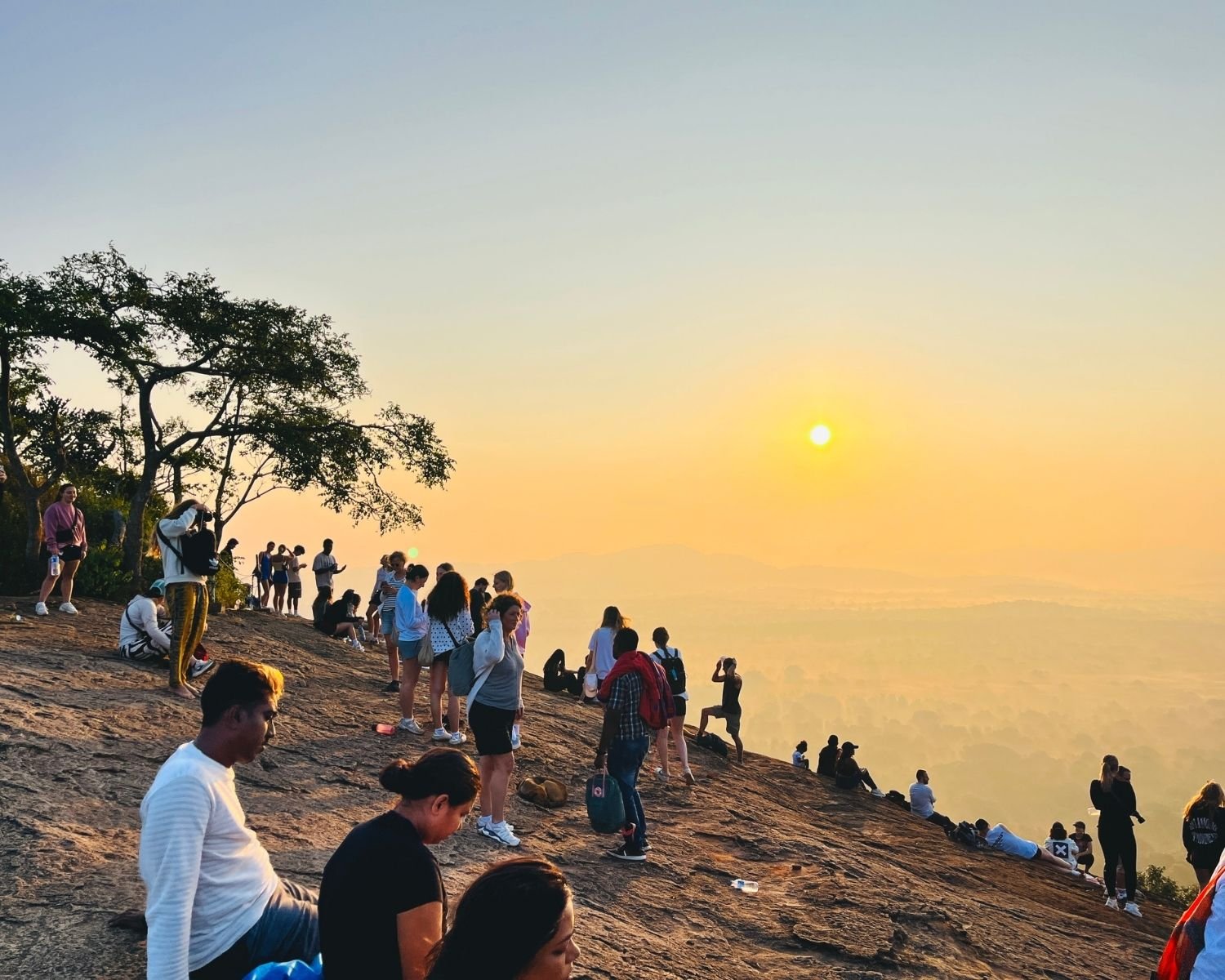 Crowds of people watching sunrise from rocky summit with tree and misty valley views at Pidurangala Rock near Sigiriya