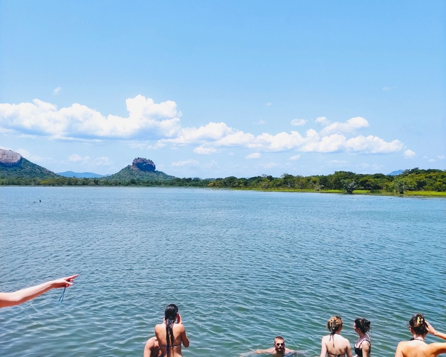 People swimming in Thalkote Wewa ancient irrigation reservoir with Lion Rock centered and Pidurangala Rock visible left across the water under blue sky near Sigiriya