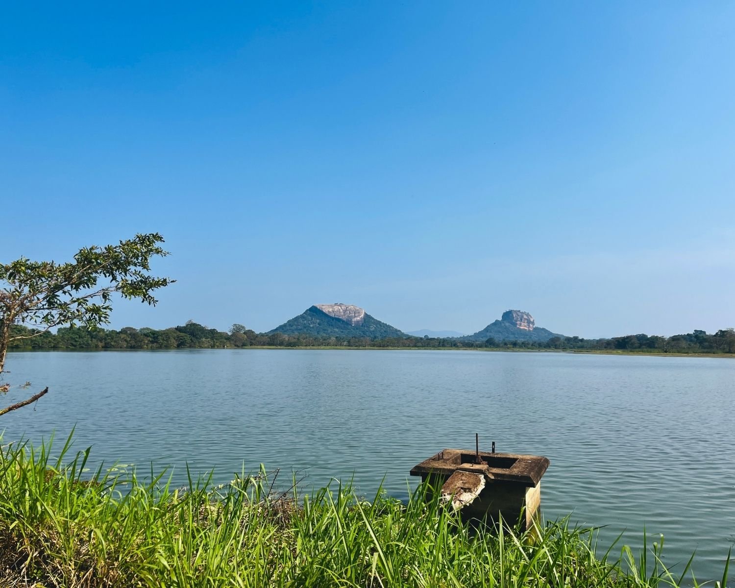 Calm Thalkote Wewa ancient irrigation reservoir with Lion Rock and Pidurangala Rock across the water, concrete sluice outlet in foreground near Sigiriya