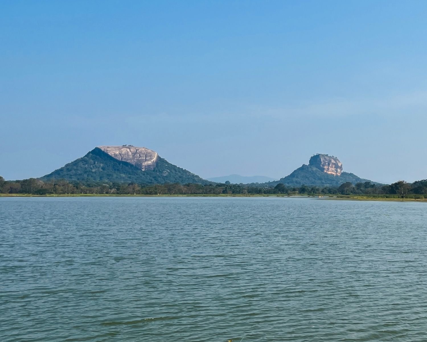 Thalkote Wewa ancient irrigation reservoir with Pidurangala Rock left and Lion Rock right across calm water under blue sky near Sigiriya