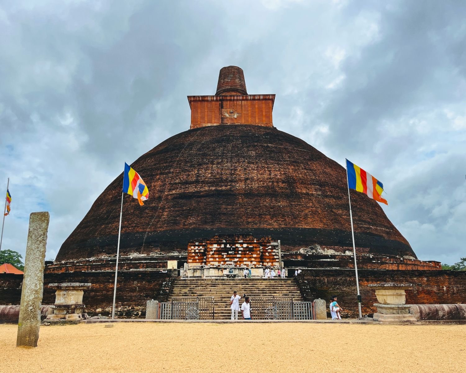 Full south view of Jetavanaramaya brick stupa with square harmika, vahalkada entrance, stone steps, and Buddhist flags
