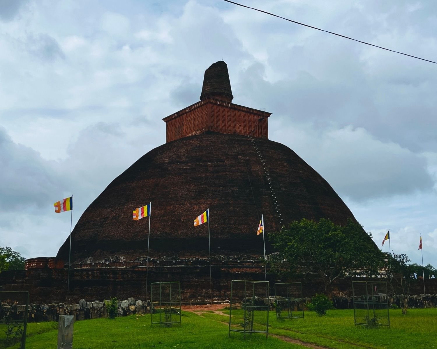 Jetavanaramaya brick stupa from northeast with square harmika, Buddhist flags, and grass parkland under cloudy sky