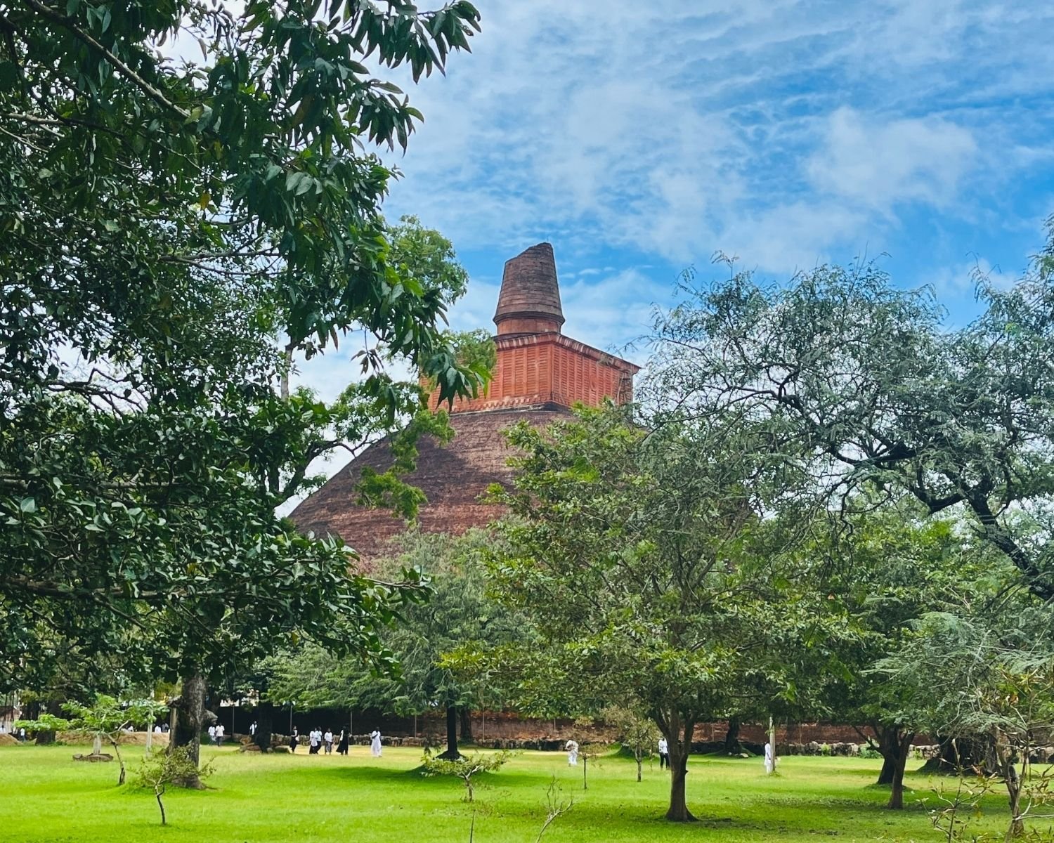Jetavanaramaya brick stupa from northeast framed by trees in the Archaeological Park with devotees on lawn