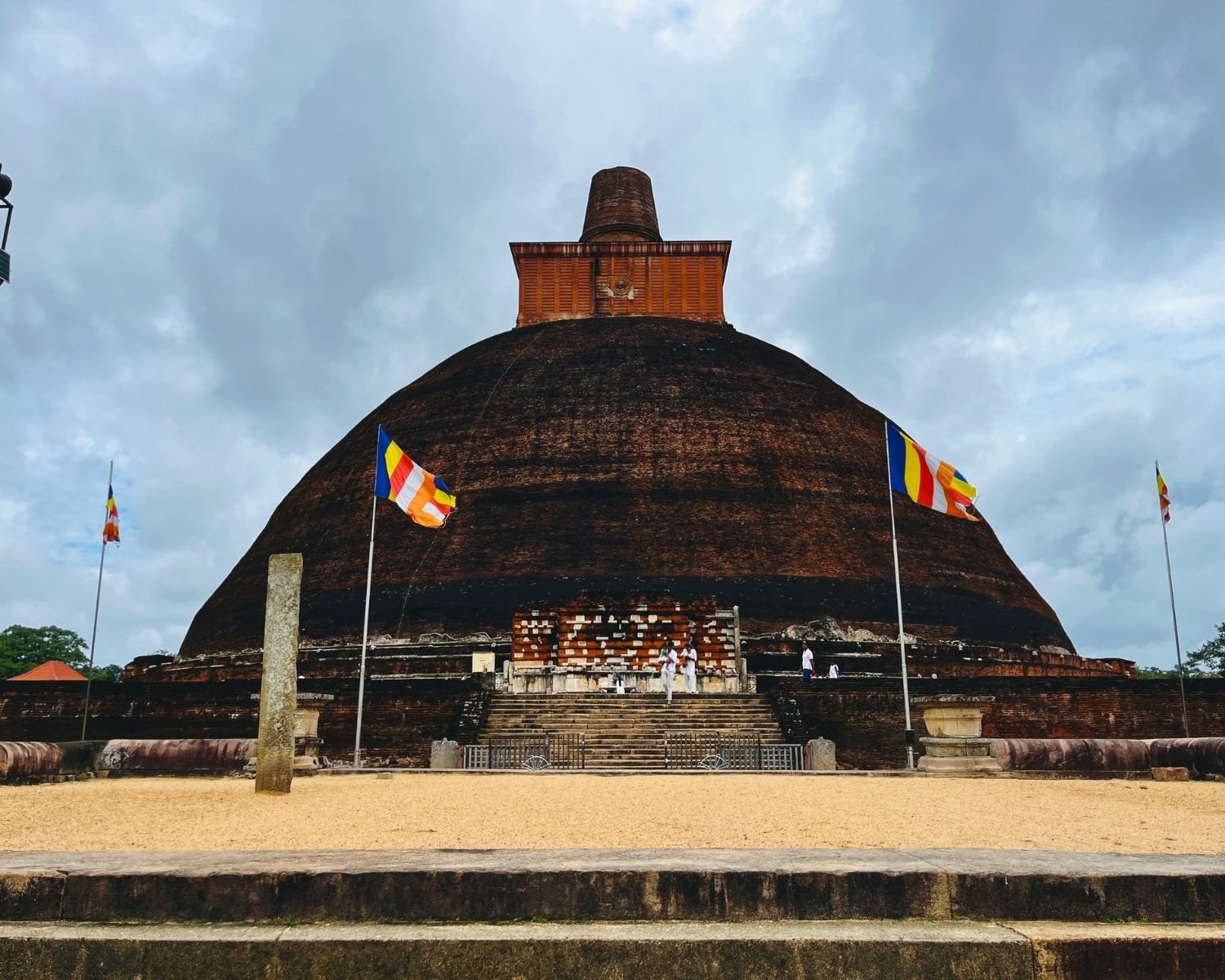 Jetavanaramaya brick stupa south entrance with stone steps, ancient pillar, and Buddhist flags under cloudy sky