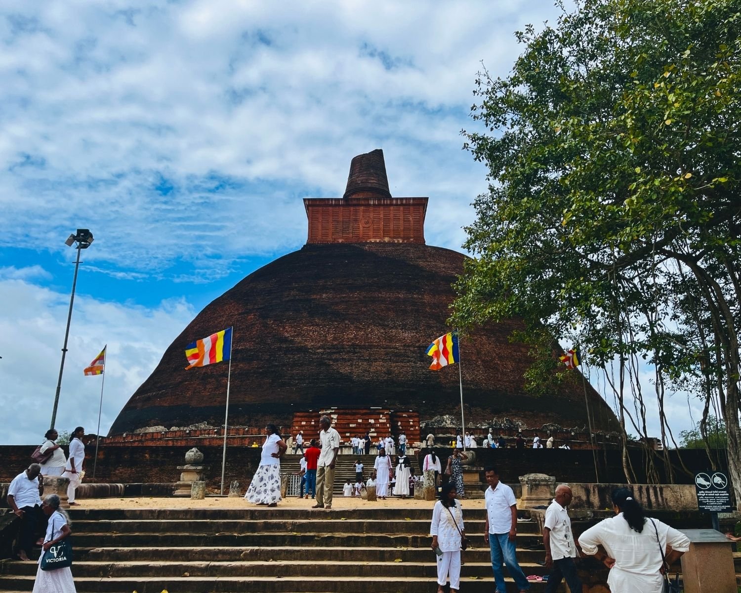 Jetavanaramaya west entrance with devotees on stone steps, Buddhist flags, and Bodhi tree beside brick stupa