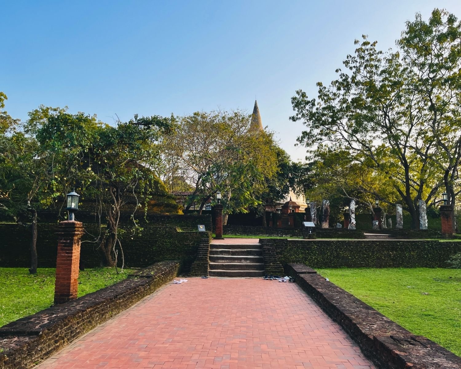 Brick causeway and steps leading to Kiri Vehera dagoba with mossy walls and lantern posts through trees