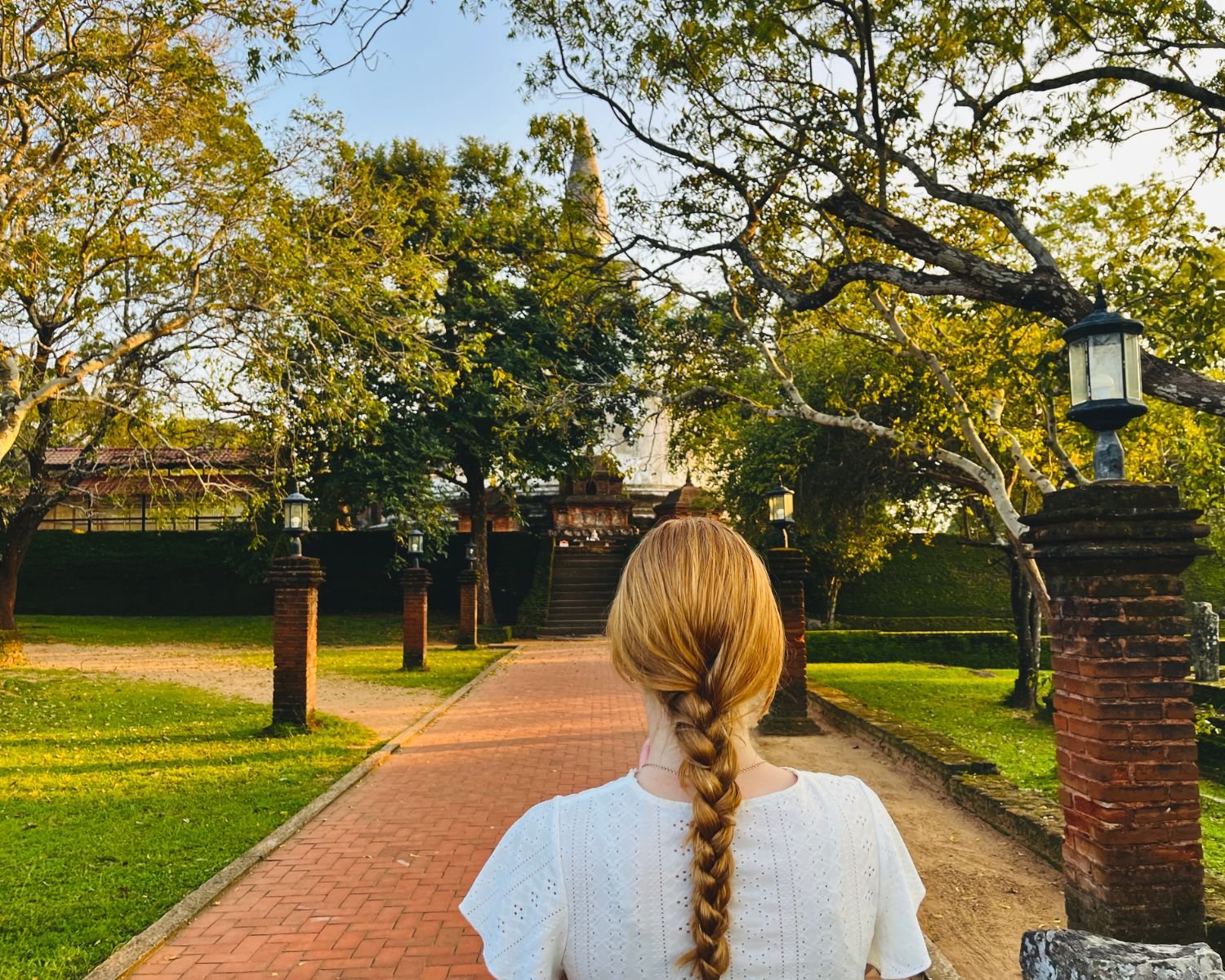 Red brick walkway with lamp posts and trees leading to staircase of Kiri Vehera stupa at sunset
