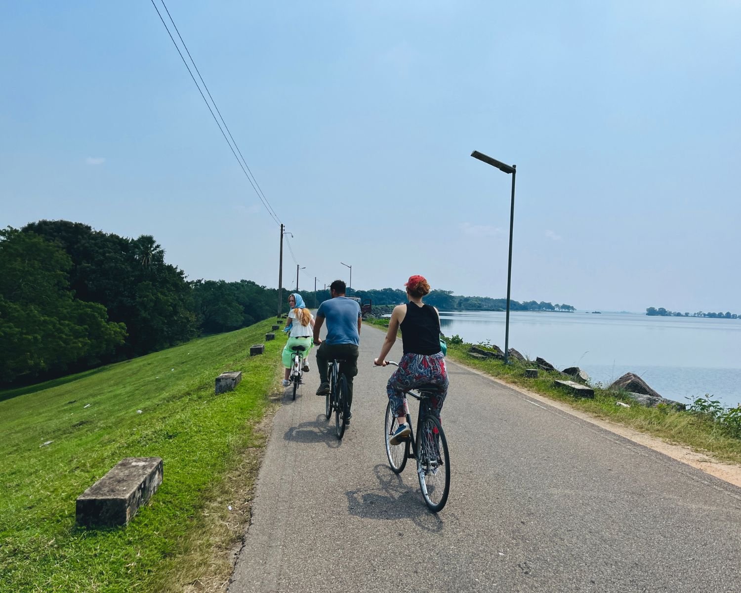 Cyclists riding along Parakrama Samudraya reservoir road beside calm water and green embankments