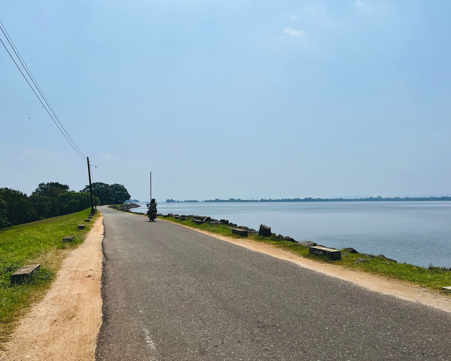 Quiet lakeside road beside Parakrama Samudraya with calm water, grass banks, and distant tree lines