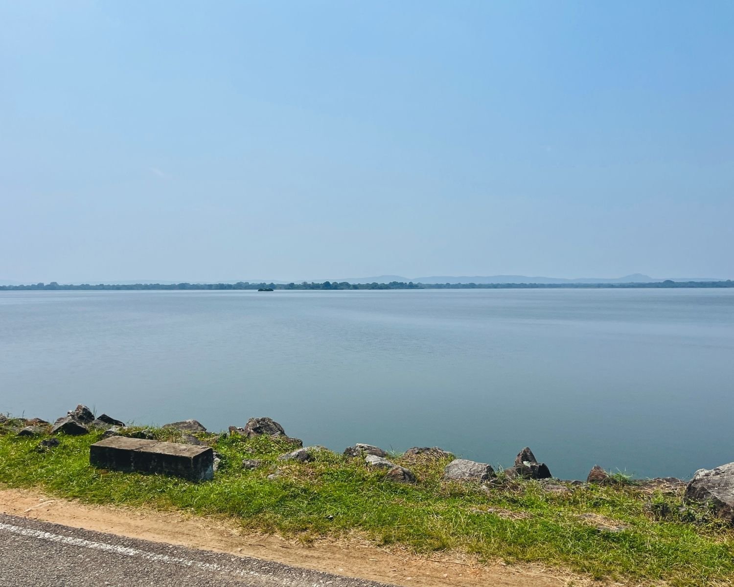 Wide open water view of Parakrama Samudraya from roadside with rocks, grass, and distant hills