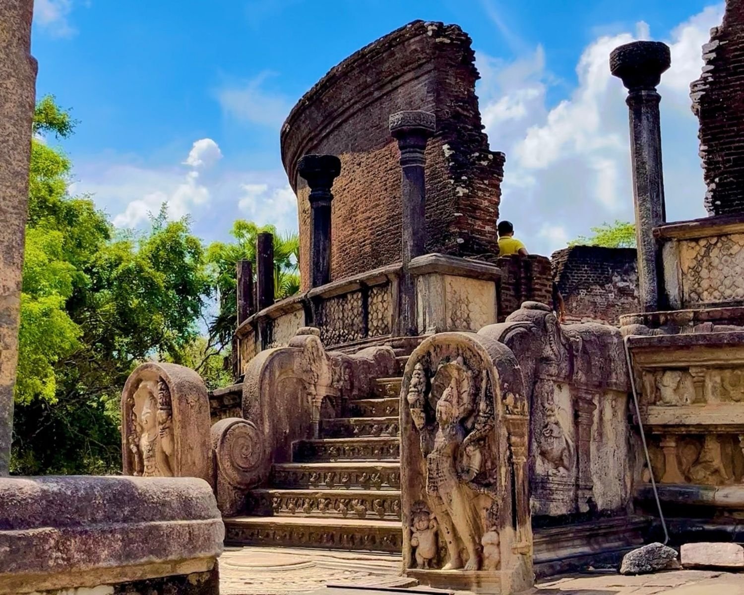 Angled view of Vatadage showing curved brick wall, stone columns, carved guard stone, and Buddha