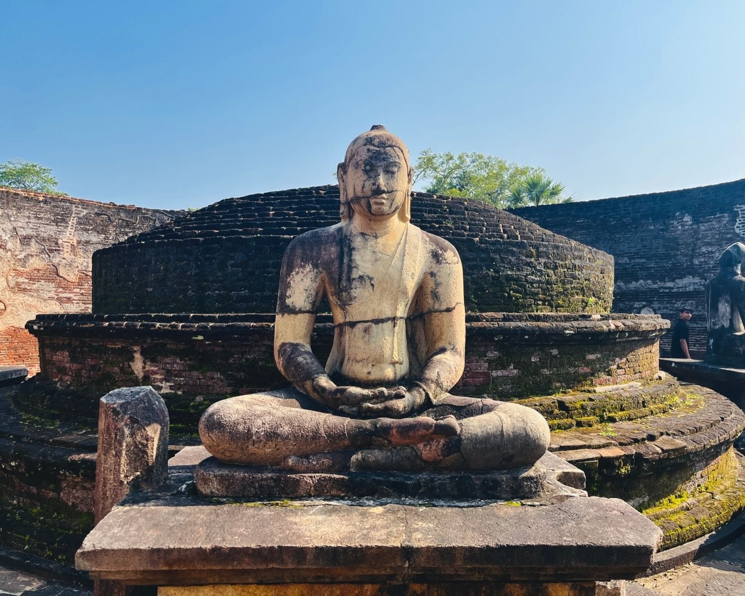 Wider view of Vatadage Buddha with mossy stupa tiers, stone pillar, and outer circular wall