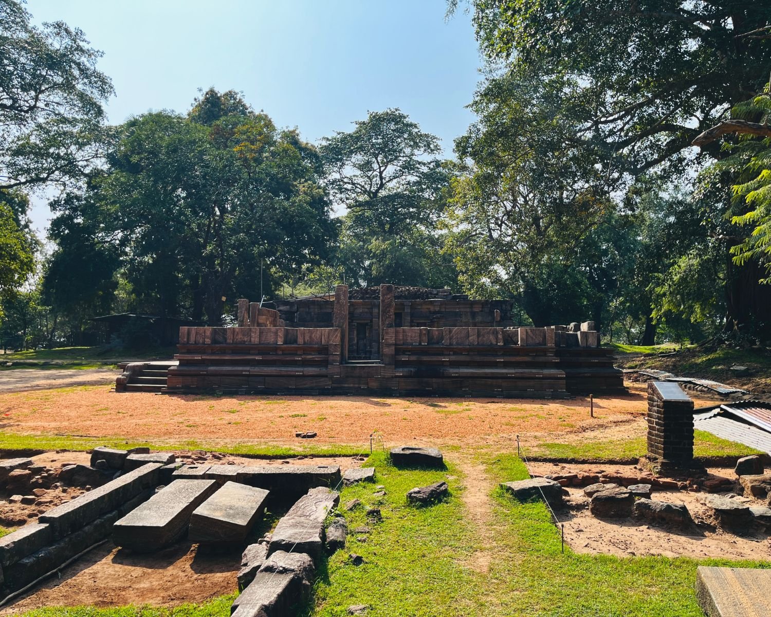 Wide front view of Shiva Devalaya No.1 showing raised stone platform, tiered walls, main entrance, and excavation trenches