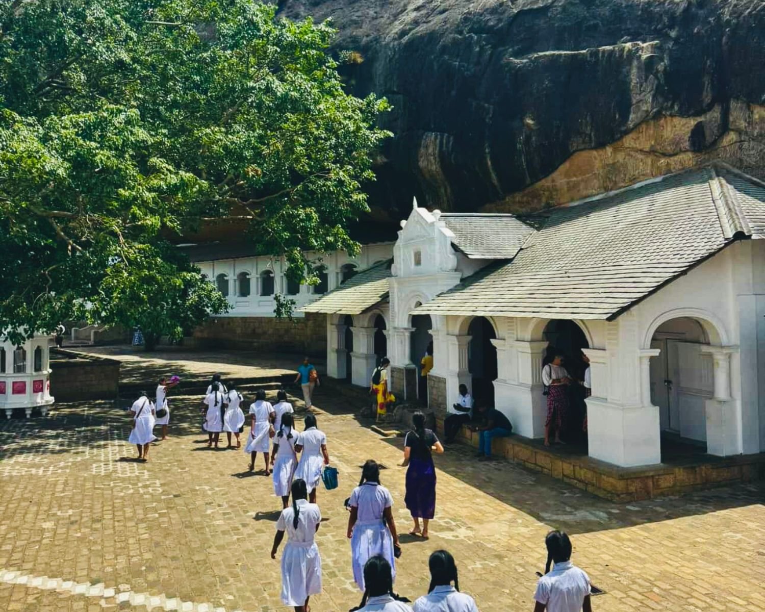 White shrine façades beneath the ancient rock shelter with visitors walking through the courtyard