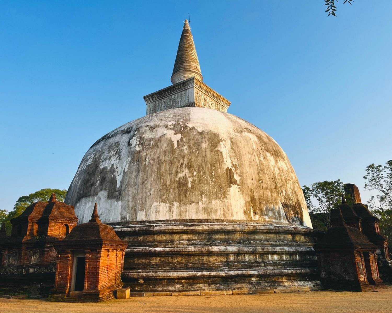White dome of Kiri Vehera stupa with spire and brick satellite shrines at base in evening light
