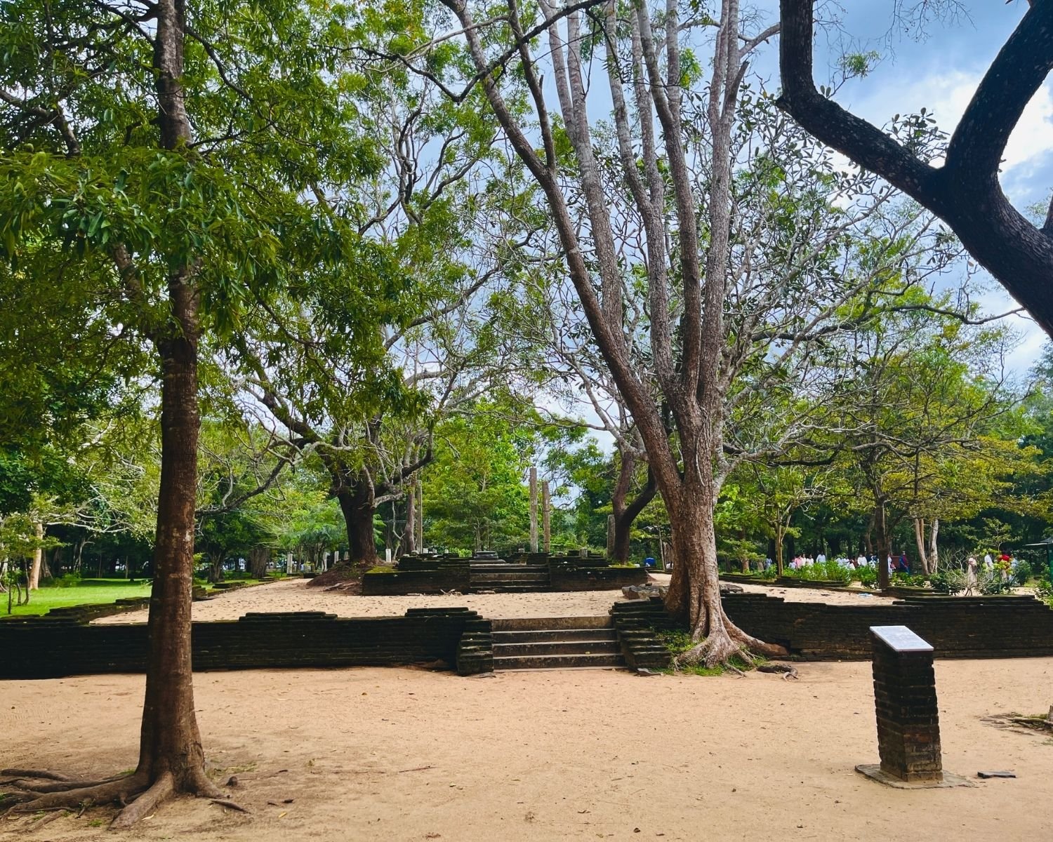 Ancient Bodhisattva Image House ruins with weathered stone steps, brick platforms, and tall stone pillars under large sacred trees, with information plaque in sandy courtyard and pilgrims visible in background