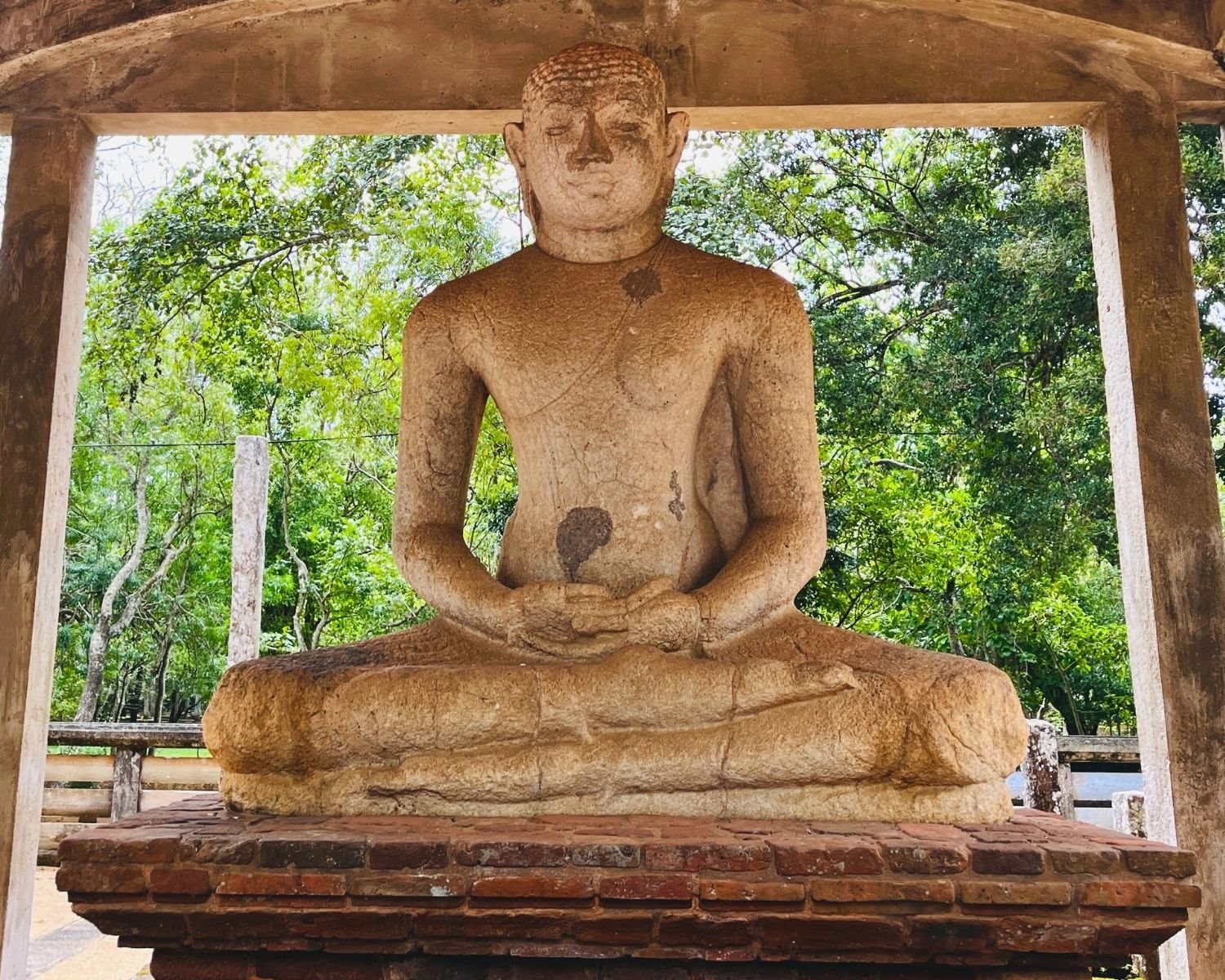 Ancient stone Samadhi Buddha statue in dhyana mudra meditation pose under stone pavilion with weathered surface, seated on brick pedestal, framed by stone pillars with lush green forest background