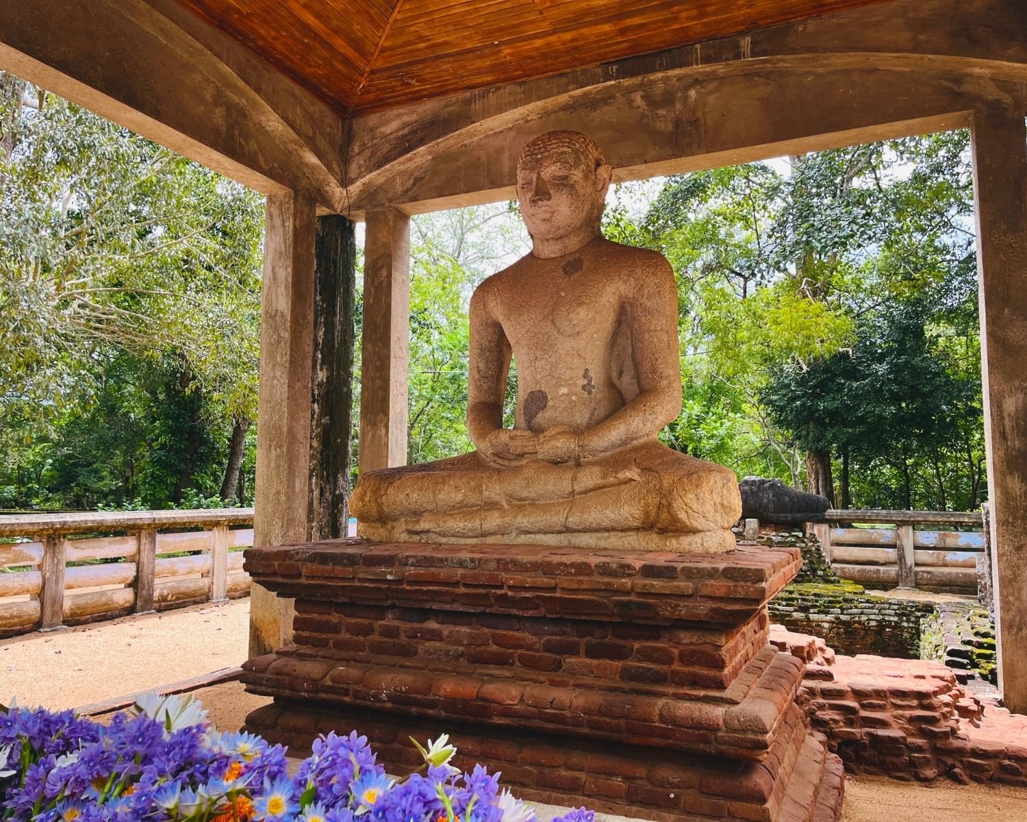 Ancient Samadhi Buddha statue in dhyana mudra under wooden-roof pavilion on layered brick pedestal, with purple and white lotus flower offerings in foreground, stone ruins and green trees behind