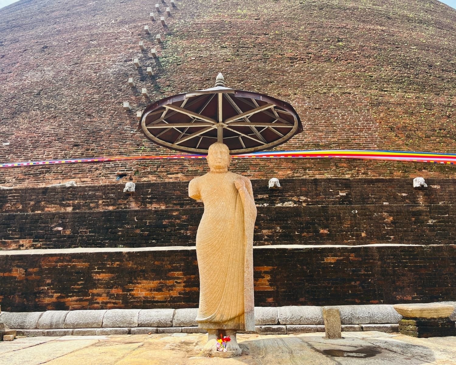 Standing stone Buddha statue under ceremonial umbrella at Abhayagiri Stupa, massive ancient brick dome with elephant carvings and colored cloth behind, flower offerings at feet in Anuradhapura