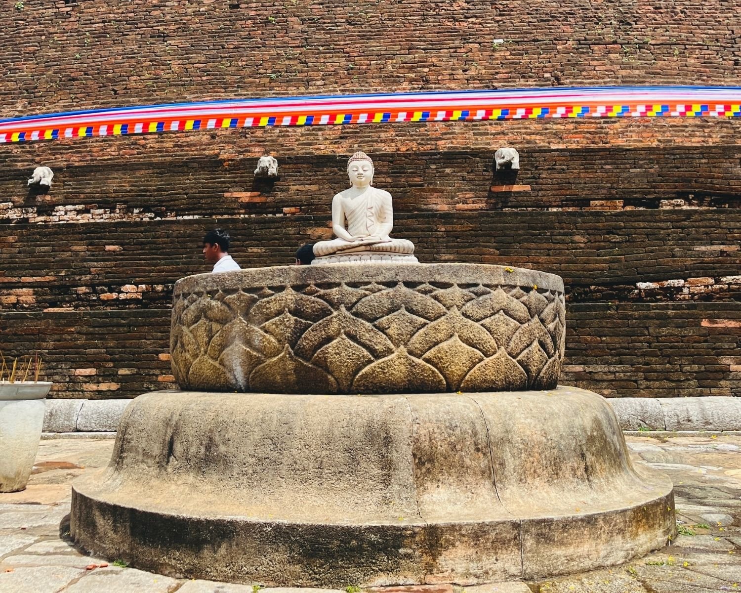 Ancient stone lotus altar carved with petal pattern at Abhayagiri Stupa, modern white Buddha statue seated on top, brick wall with elephant waterspouts and ceremonial cloth behind in Anuradhapura