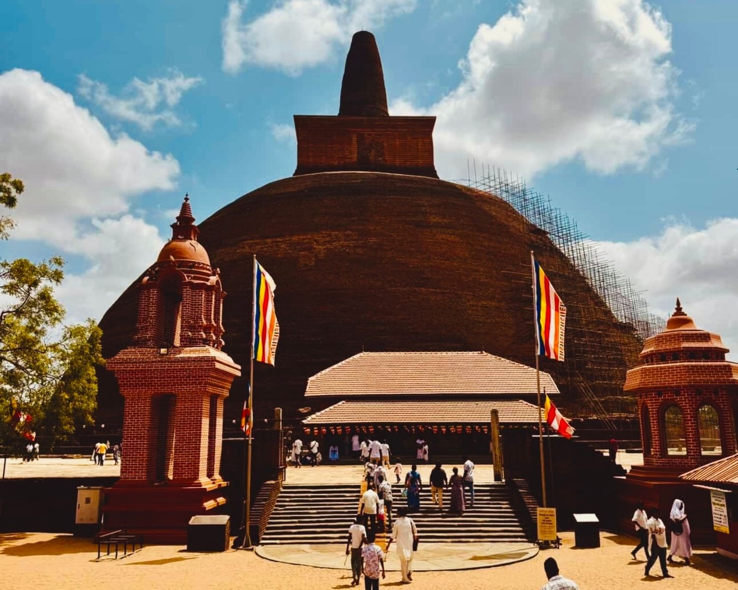 Abhayagiri Stupa massive exposed brick dome with pointed conical spire and square harmika, twin brick shrines, Buddhist flags, pilgrims on stairs and scaffolding under blue sky at Anuradhapura