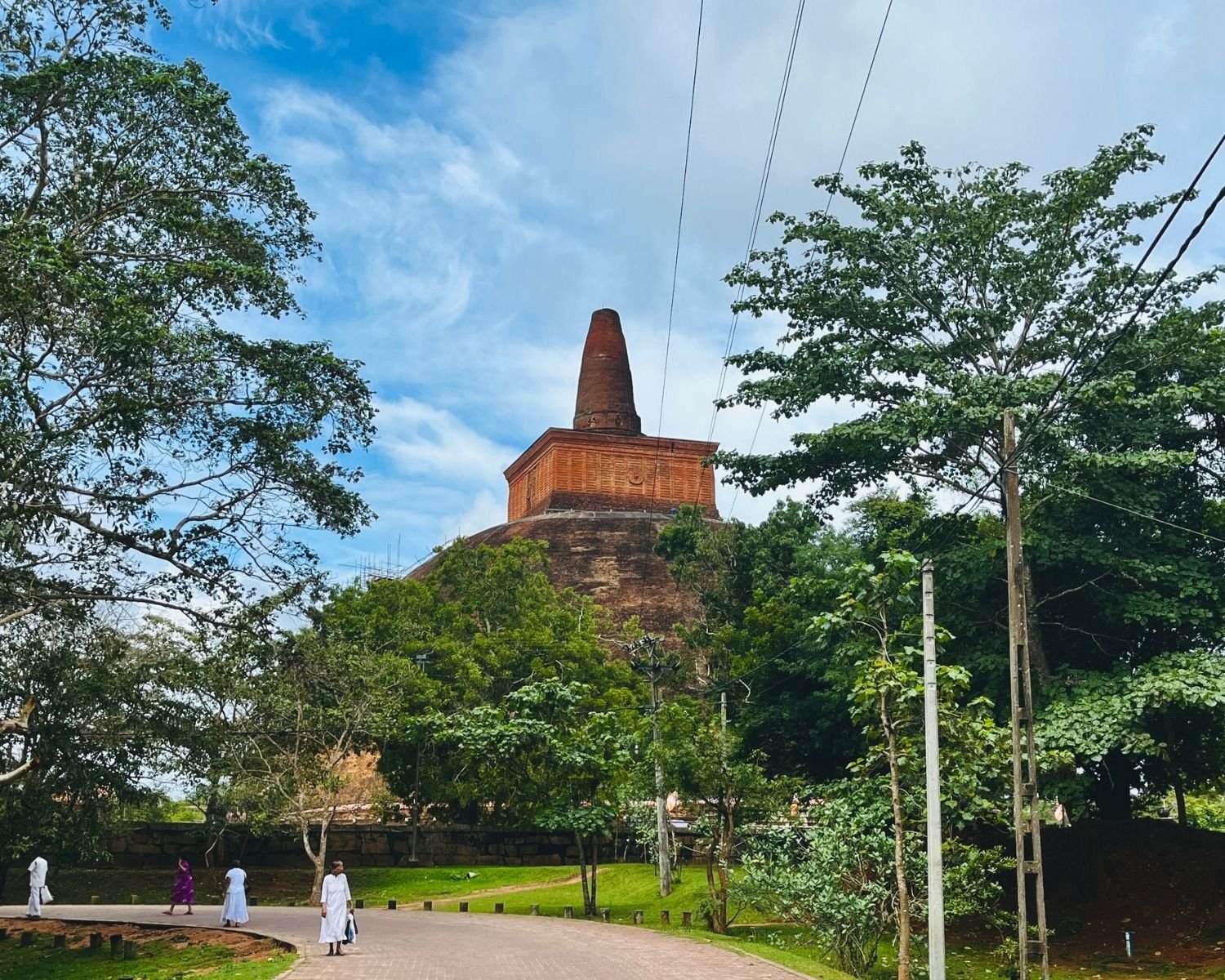 Abhayagiri Stupa brick dome, square harmika and conical spire seen from east approach path, framed by trees with pilgrims walking toward stupa, scaffolding left, power lines overhead in Anuradhapura