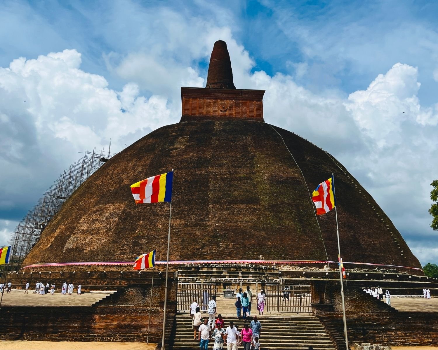 Abhayagiri Stupa east face showing massive brick dome, square harmika with medallion and conical spire, bamboo scaffolding, stone stairway and Buddhist flags under clouds in Anuradhapura
