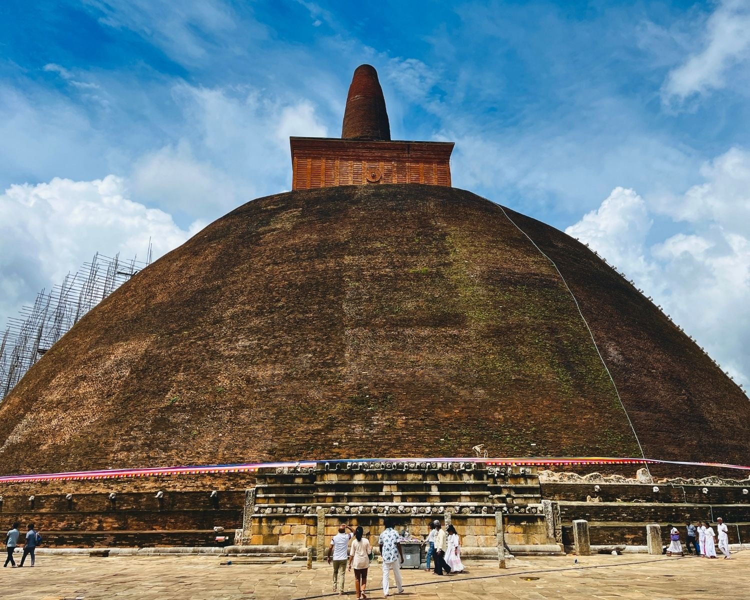 Close view of Abhayagiri Stupa east face showing massive weathered brick dome, square harmika with circular medallion, conical spire, bamboo scaffolding and stone platform with devotees in Anuradhapura