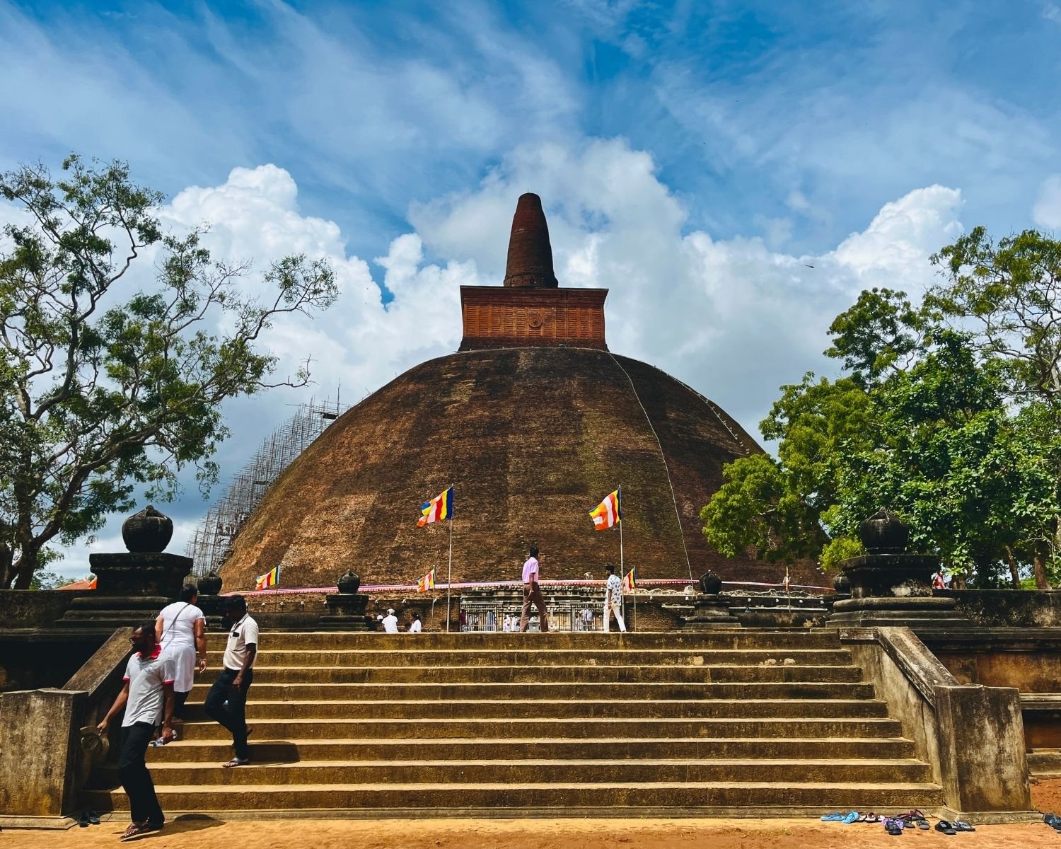 Abhayagiri Stupa east face with massive exposed brick dome, square harmika and conical spire, stone stairway, metal gate, Buddhist flags and bamboo scaffolding under cloudy sky in Anuradhapura